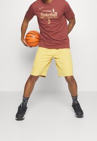 Punainen Nike t-paita, jossa on kultainen NBA-logo, keltaiset shortsit, mustat lenkkarit ja kuviolliset mustat sukat, holdinga oranssia koripalloa.