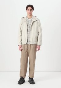 Lichtbeige zip-up jas met capuchon, over een gestreept shirt, gecombineerd met losse tankleurige broek en donkere schoenen. Gladde textuur, casual pasvorm.