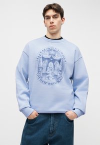 Nuori mies, jolla on korvakorut, pukeutunut vaaleansiniseen svetariin, jossa on pyöreä kuva, jossa hahmoja ja leijona, sekä teksti "THE NEW ORIGINALS."