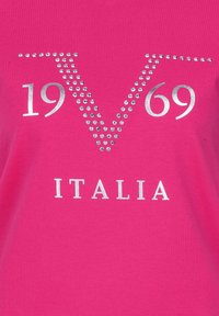 Pink T-shirt featuring a silver, rhinestone-studded “V” design with “1969” and “ITALIA” printed underneath in silver. Soft fabric texture.
