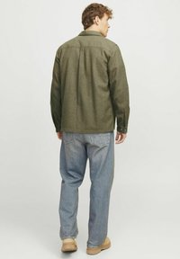 Zeleno, teksturirano flanelasto srajco z yoke na rami, gumbi na manšetah in hrbtno plisirano, v kombinaciji z svetlo modrimi jeans hlačami in bež obutvijo.