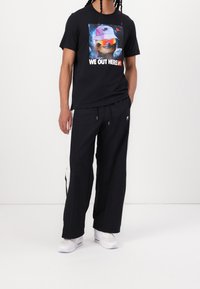 Person som bär svarta byxor med vita ränder, vita sneakers och en svart t-shirt med en sengångare i rymden och texten "VI ÄR HÄR".