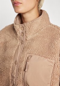 Beige fleecejakke med en blød, shaggy tekstur, trykknapper og en lynlåslomme på forsiden. Kravedesignet tilføjer varme.
