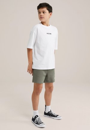 T-shirt en coton blanc avec du texte noir, associé à un short vert olive. Des baskets noires et blanches et des chaussettes blanches complètent la tenue.