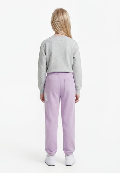 Hosszú szőke hajú gyermek áll háttal, szürke hosszú ujjú felsőt, világoslila melegítőnadrágot és fehér sportcipőt visel fehér háttér előtt.