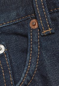 Gros plan sur un tissu denim bleu montrant un rivet en cuivre, une couture jaune et une boucle de ceinture sur un jean.