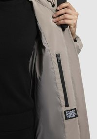 Gai beigā krāsa jaka ar iekšējo rāvējslēdzēja kabatu un birku, ar gludu, matētu tekstūru. Ārpusei ir vienkāršs, plūstošs dizains.