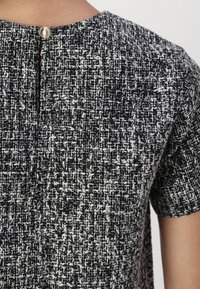 Tessuto texturizzato in bianco e nero con un motivo tessuto e maniche corte; presenta una chiusura sul retro con un bottone metallico rotondo.