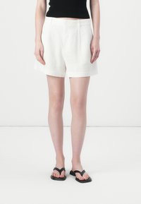 Pantaloni corti bianchi in lino con un design su misura, caratterizzati da una vita media e orli arrotolati, abbinati a infradito nere.