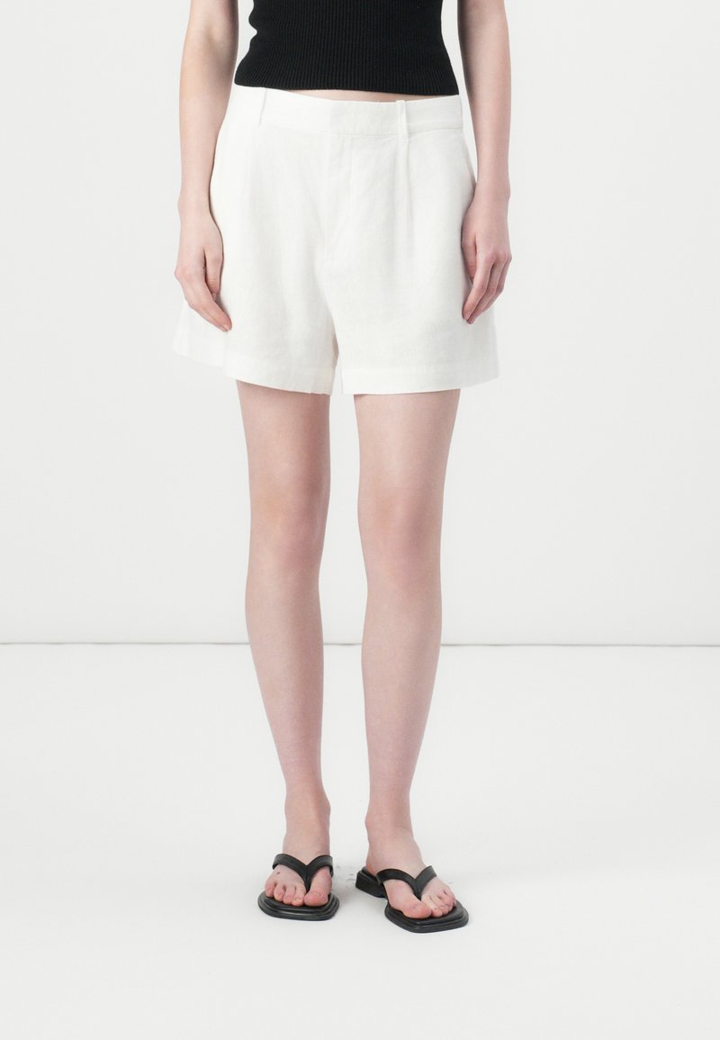 Pantaloni corti bianchi in lino con un design su misura, caratterizzati da una vita media e orli arrotolati, abbinati a infradito nere.