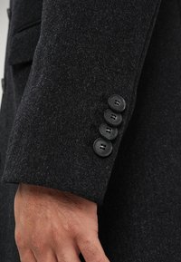 Polsino di un cappotto di lana nera con quattro bottoni neri, tessuto texturizzato e vestibilità su misura. Il polsino presenta un dettaglio a notche vicino al polso.