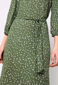 Robe verte avec un motif abstrait blanc en forme de graines, manches longues et ceinture en tissu nouée à la taille, montrée de la moitié du torse jusqu'aux mains.