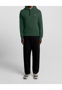 Felpa verde con cappuccio regolabile, tasca a marsupio e un piccolo logo dorato. Abbinata a pantaloni a gamba larga neri e sneakers grigie.