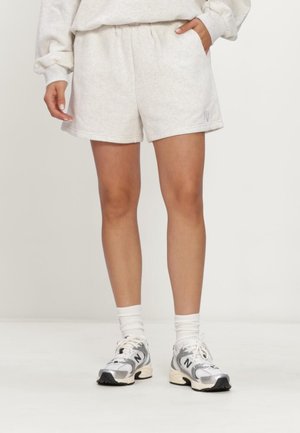 Persoon draagt lichtgrijze casual shorts, witte sportsokken en witte en grijze New Balance sneakers, staand met één hand in de zak.