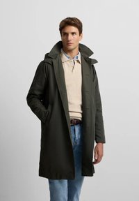 Abrigo impermeable de color verde oscuro con un gran collar, botones frontales y bolsillos laterales, llevado sobre un polo beige y unos vaqueros azul claro.