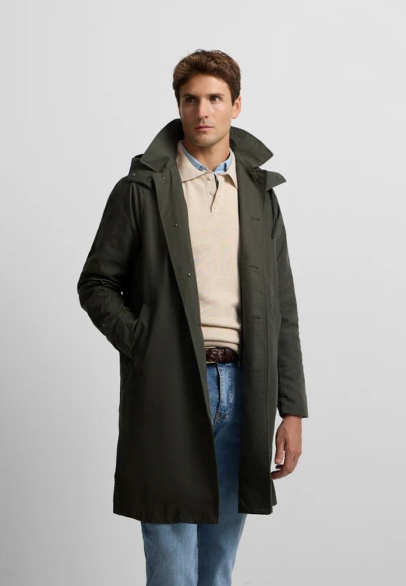 Abrigo impermeable de color verde oscuro con un gran collar, botones frontales y bolsillos laterales, llevado sobre un polo beige y unos vaqueros azul claro.