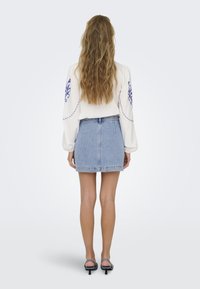 White long-sleeve shirt with blue embroidery, paired with a light blue denim skirt. Model wearing black heeled sandals; wavy hair.