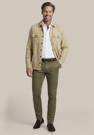 Lachende man staand, draagt een beige jas met knopen, wit overhemd, olijfgroene broek, bruine gevlochten riem en donkerbruine instapschoenen.