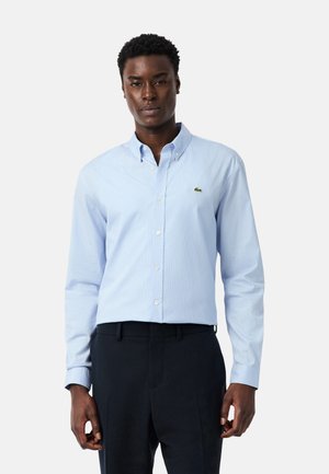 Homme portant une chemise bleu clair boutonnée avec un petit logo de crocodile vert sur la poitrine, associée à un pantalon sombre.