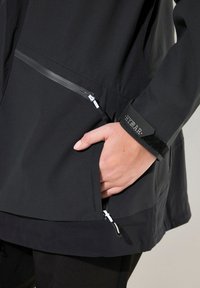 Chaqueta negra impermeable con un bolsillo con cremallera, tejido texturizado y puños ajustables de velcro para un ajuste seguro.