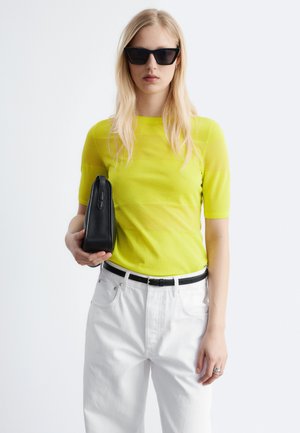 Femme aux longs cheveux blonds portant des lunettes de soleil noires, un haut à manches courtes jaune, un pantalon blanc, une ceinture noire et tenant une pochette noire.