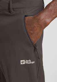 Dunkelgraue Outdoor-Hosen mit einer Reißverschlusstasche, elastischem Kordelzug und sichtbarem Logo auf dem Oberschenkel. Glatte Textur und taillierte Passform.