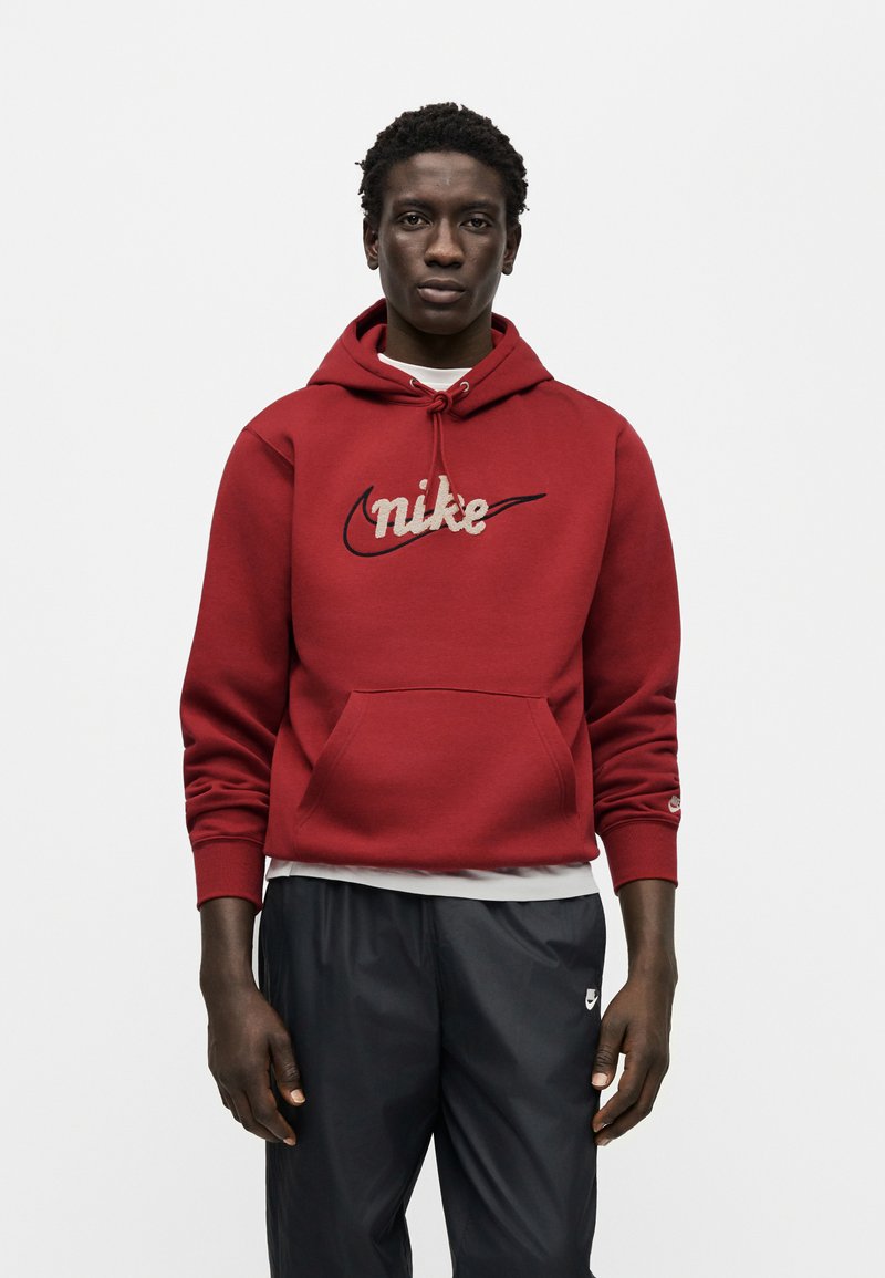 Felpa con cappuccio Nike rossa con tasca a marsupio frontale, logo ricamato beige, cappuccio con coulisse e polsini a coste, abbinata a pantaloni sportivi neri.