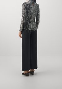 Schwarz-silbernes Paillettenoberteil mit hohem Kragen und Schlüssellochausschnitt am Rücken, kombiniert mit weit geschnittenen schwarzen Hosen und braunen Keilschuhen.