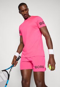 Rosa idretts t-skjorte og shorts med "BORG" merke, teksturert stoff, tettsittende design, og en tennisball i den ene hånden, racket i den andre.
