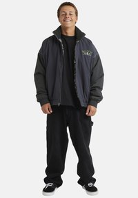 Black and gray oversized jacket with a high collar, zip closure, and embroidered logo, paired with black baggy pants and black sneakers.
