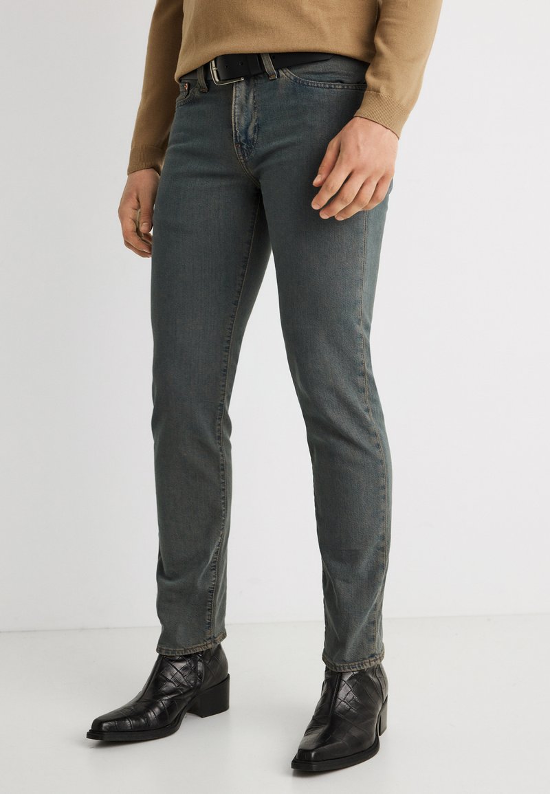 Άνδρας που φοράει ξεθωριασμένο σκούρο γκρι τζιν στενής γραμμής, μαύρα δερμάτινα μποτάκια με υφή στον αστράγαλο και μπεζ πουλόβερ με μακριά μανίκια, στέκεται μπροστά σε λευκό φόντο.