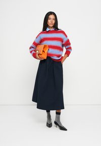 Maglione a righe rosso e blu, gonna a pieghe blu navy, calzini grigi, tacchi neri, mentre tiene una pochette arancione. Sfondo semplice.