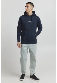 Navy blue hoodie with a pocket, featuring a white graphic design. Paired with light blue jeans and black sneakers. Casual fit and style.