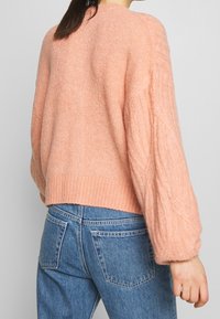 Rosa stickad tröja med avslappnad passform, ribbat avslut och flätstickade ärmar, parat med blåjeans. Texturen verkar mjuk och fluffig.