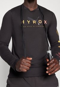 Maglietta atletica a maniche lunghe nera realizzata in tessuto elasticizzato, con logo in testo arancione e giallo. Indossata con una corda per saltare in mano.