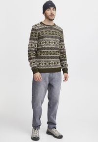 Strickpullover in olivgrün mit geometrischen Mustern in Grau und Creme. Getragen mit grauen Jeans und grauen Turnschuhen, ergänzt durch eine dunkle Mütze.