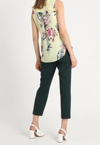 Blusa senza maniche floreale verde chiaro con motivi rosa e viola, abbinata a pantaloni aderenti verde scuro e sandali bianchi con tacco grosso.