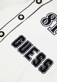 Baltas džersis švarkas su juodais mygtukais, kontrastine siuvinėjimo linija ir išsiuvinėtu "GUESS" logotipu per krūtinę.
