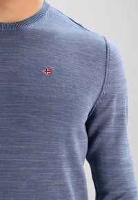 Maglione blu lavorato a maglia con scollo rotondo e una piccola bandiera norvegese ricamata sul petto. Tessuto liscio con un leggero motivo melangiato.
