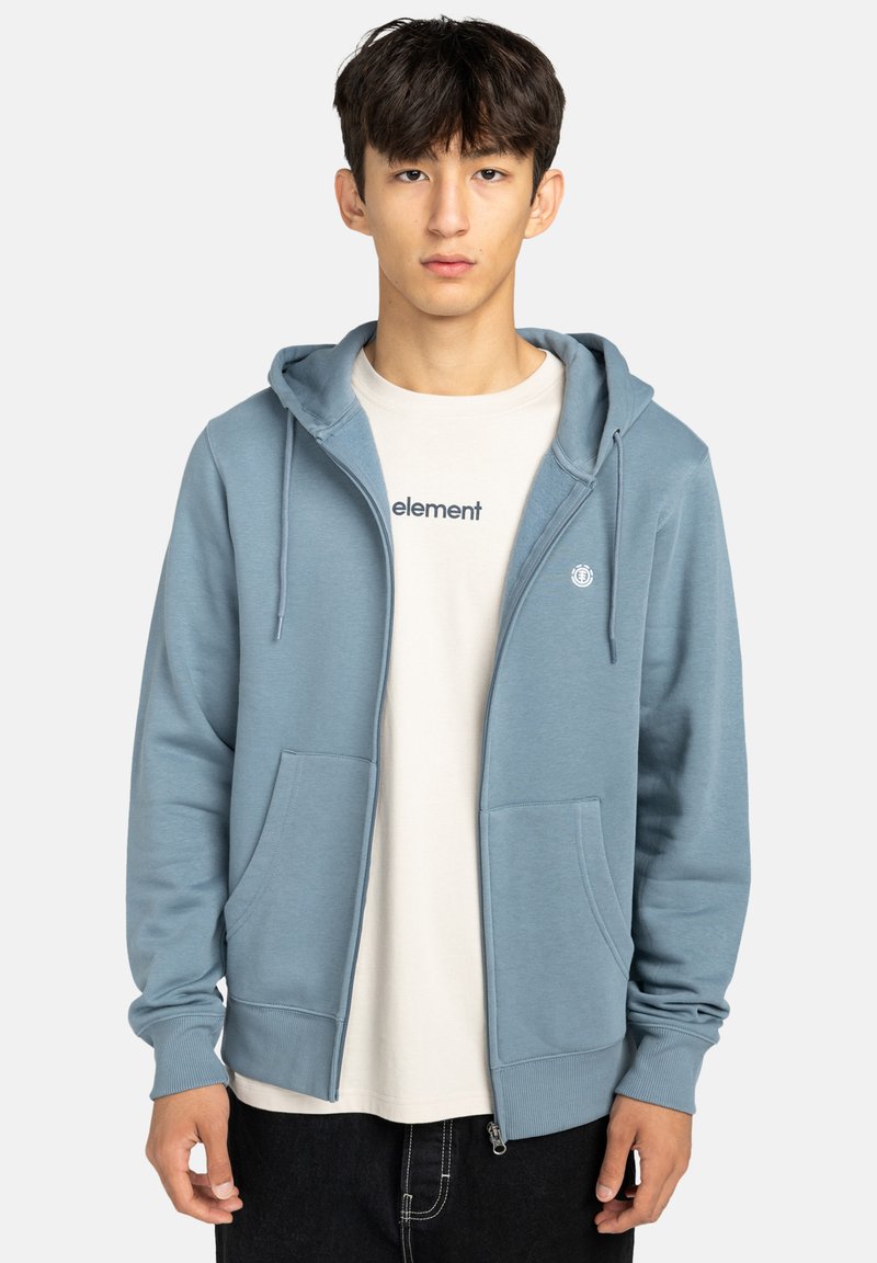 Felpa zipata azzurro chiaro con tasca a marsupio, polsini e orlo a costine, indossata sopra una t-shirt beige con il testo "element".
