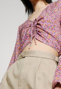 Top corto floreale con motivi rosa, arancioni e viola; presenta una chiusura con laccio davanti e maniche lunghe. Abbinato a pantaloni beige a vita alta.