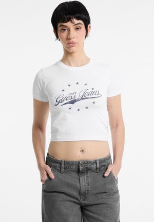 Skraťané biele tričko s grafickým motívom "Guess Jeans" v námorníckej modrej farbe, obklopené hviezdami. Spárované s vysokými sivými nohavicami.