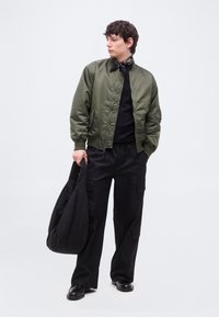 Νέος άνδρας με πράσινο bomber μπουφάν, μαύρο παντελόνι cargo, μαύρα παπούτσια και μπαντάνα στο λαιμό, κρατώντας μια μεγάλη μαύρη τσάντα, κοιτάζει προς τα αριστερά.