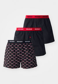 Trois paires de shorts : deux noirs avec des bandes contrastantes, rouge et marine, et un noir à motifs avec le logo répété "HUGO" en rouge et blanc.