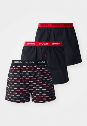 Τρία ζευγάρια σορτς: δύο μαύρα με αντίθετες λωρίδες, κόκκινη και ναυτική, και ένα μαύρο με μοτίβο που επαναλαμβάνει το λογότυπο "HUGO" σε κόκκινο και λευκό.