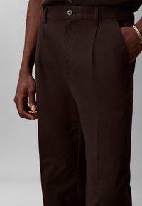 Pantalon marron foncé taillé sur mesure porté par une personne avec une main dans la poche et l'autre reposant à côté de la jambe.