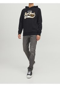 Felpa navy con logo frontale bianco e oro, abbinata a jeans grigi e sneakers nere con suola bianca e lacci neri.