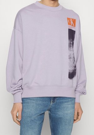 Lila mikina s černým štětcovým vzorem a oranžovým logem Calvin Klein Jeans na prsou, nošená s modrými džínami.