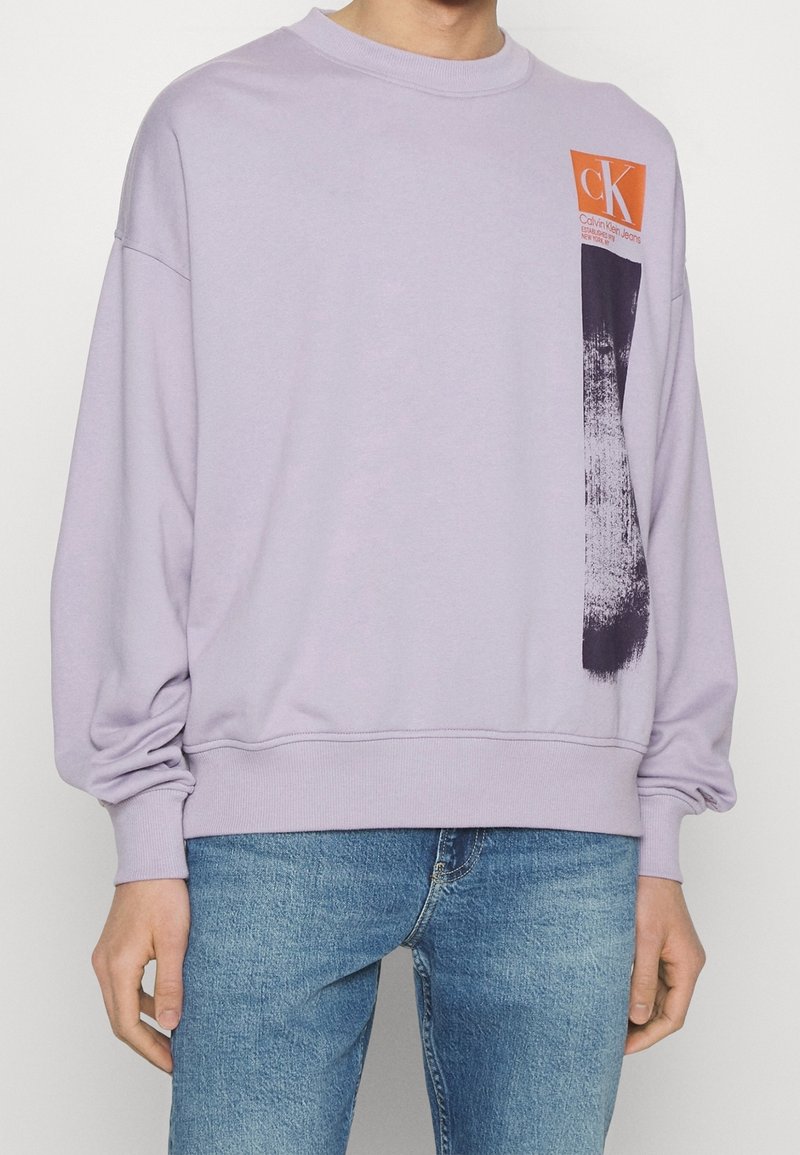 Sweatshirt en lavande avec un motif de coups de pinceau noirs et un logo Calvin Klein Jeans orange sur la poitrine, porté avec un jean bleu.
