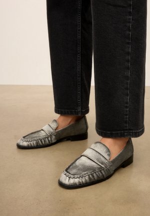 Personne portant des mocassins argentés texturés avec des semelles noires et un jean noir raccourci, debout sur un sol beige.